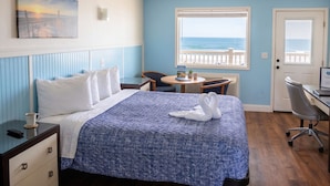 A hotel room with a bed, a desk, a chair, a window with a view of the beach, and a painting on the wall. - Blue Heron Motel (Nags Head)