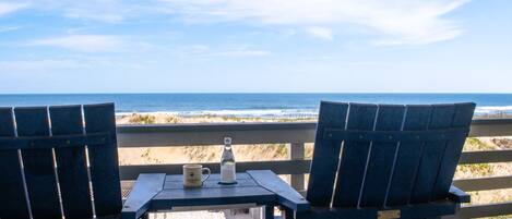 Two blue chairs with a table and a bottle on a deck overlooking the ocean.