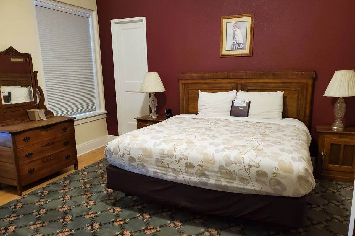 A bedroom with a floral-patterned bedspread, a wooden dresser, a nightstand with a lamp, and a framed picture on the wall.