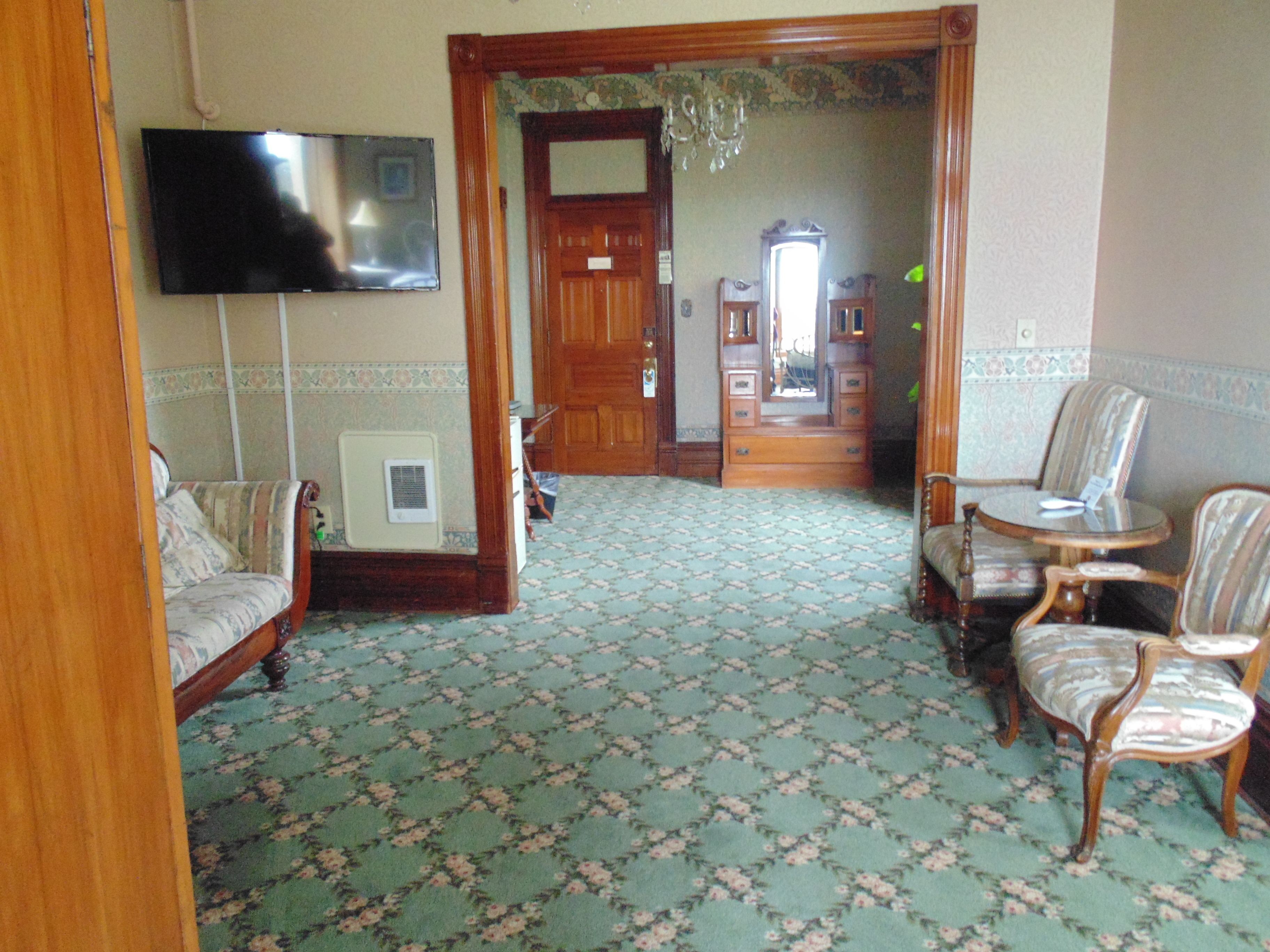 Una habitación con alfombra estampada, un sofá, una silla, una mesita y un televisor colocado en la pared.