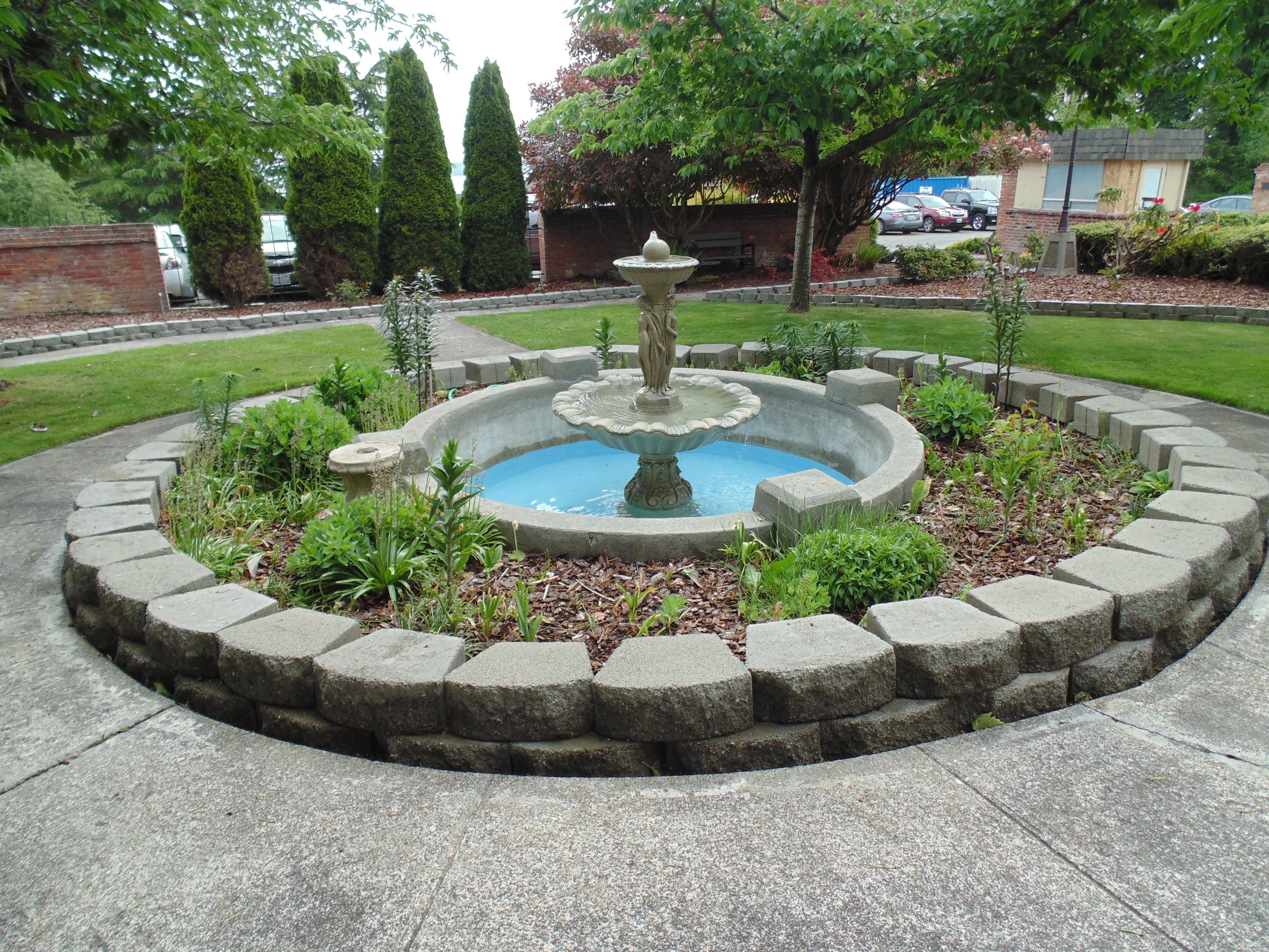 A circular stone fountain with a central statue, surrounded by a low stone border and various plants.