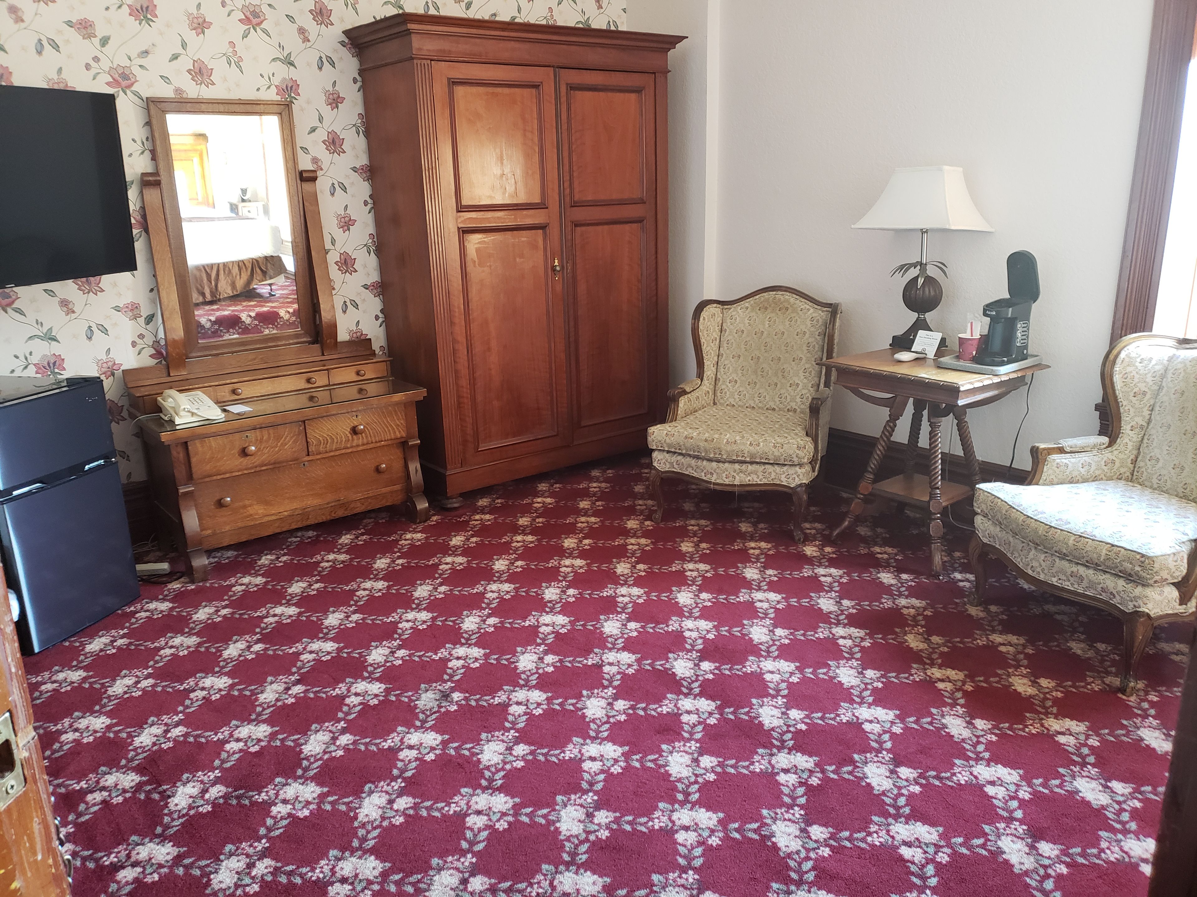 Ein Zimmer mit Holzschrank, kleinem Tisch mit Lampe, zwei Stühlen, Kommode und Fernseher.