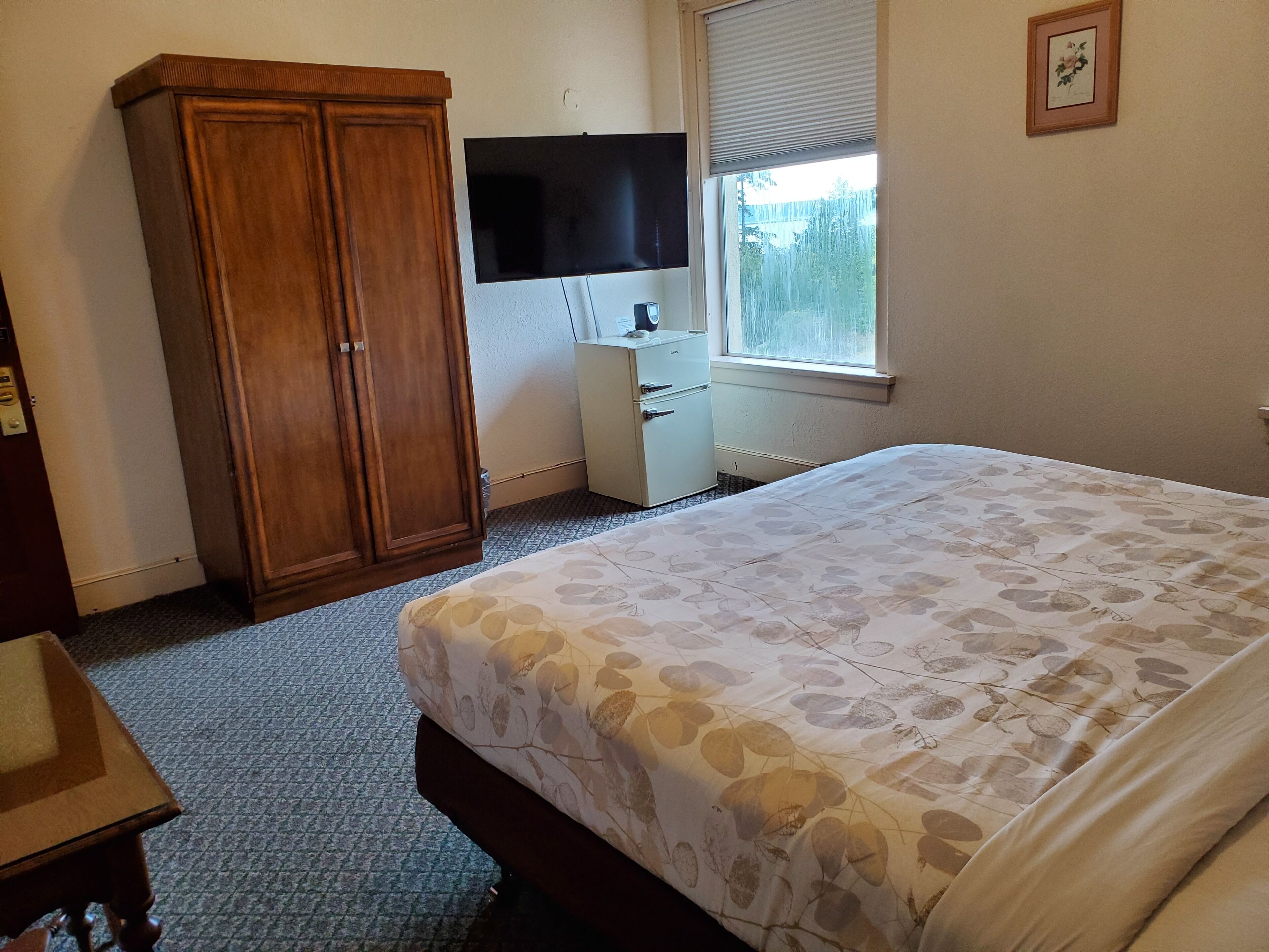 A bedroom with a bed, a wardrobe, a desk, a television, and a window with blinds.