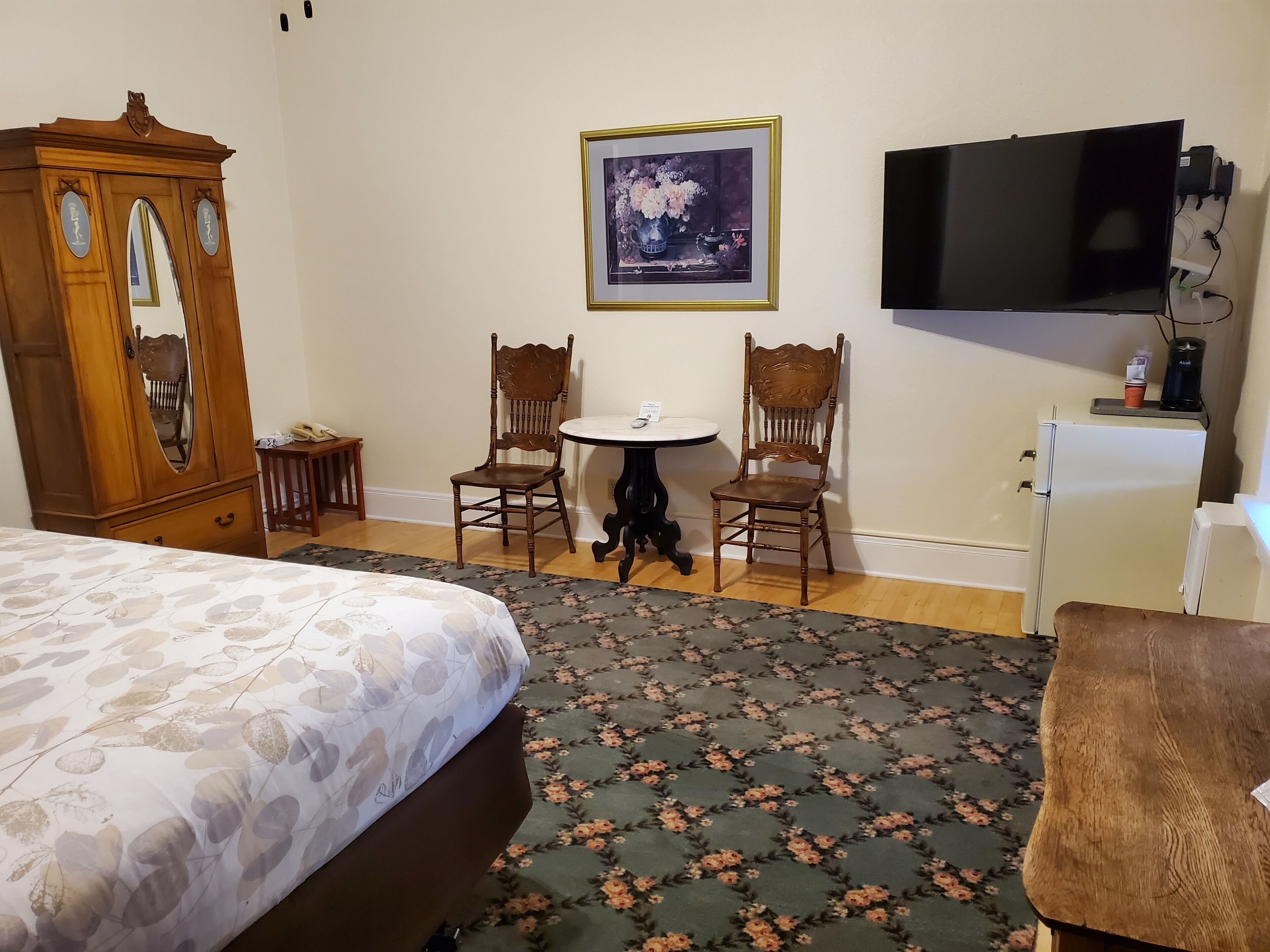 Une chambre d’hôtel comprenant un lit, deux chaises en bois, une petite table ronde, une armoire blanche, un téléviseur et un tableau encadré accroché au mur.