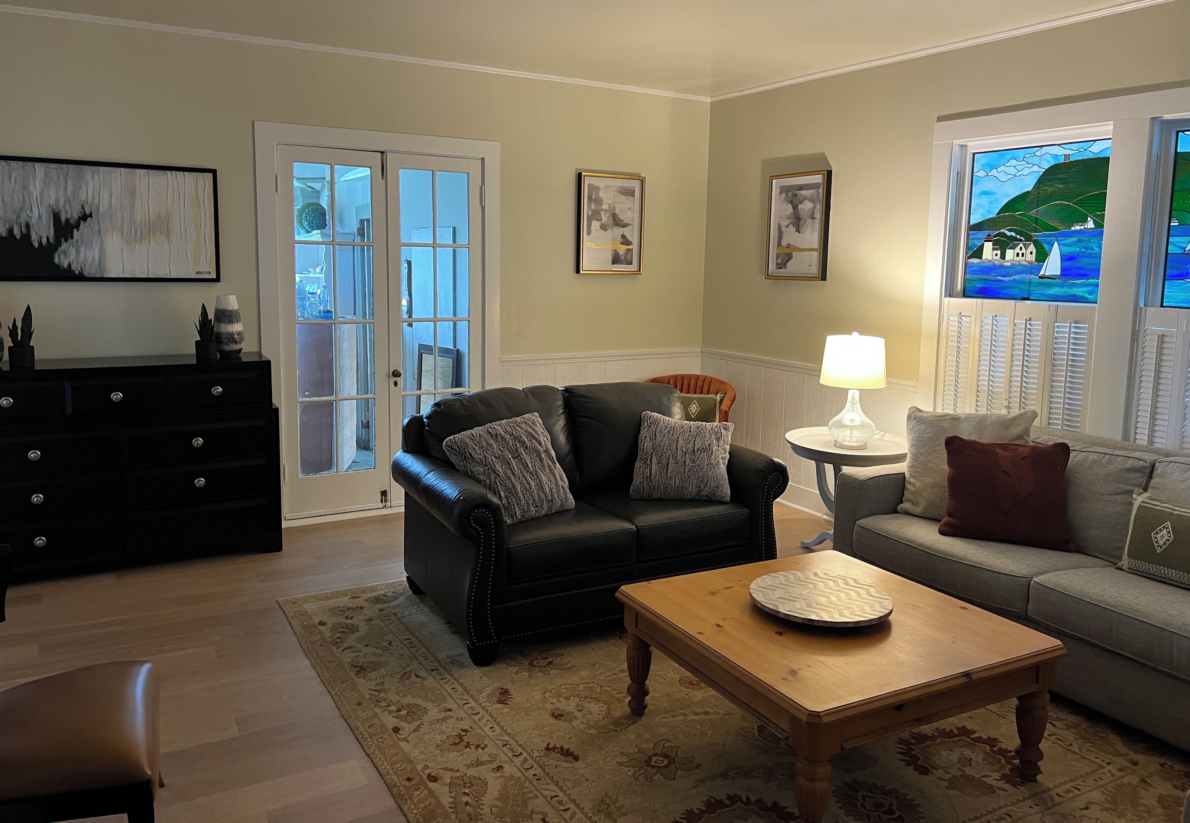 A living room with a black leather sofa, a wooden coffee table, and a grey sofa. There is a black dresser with decorative items, a patterned rug, and a window with a view of the sea.