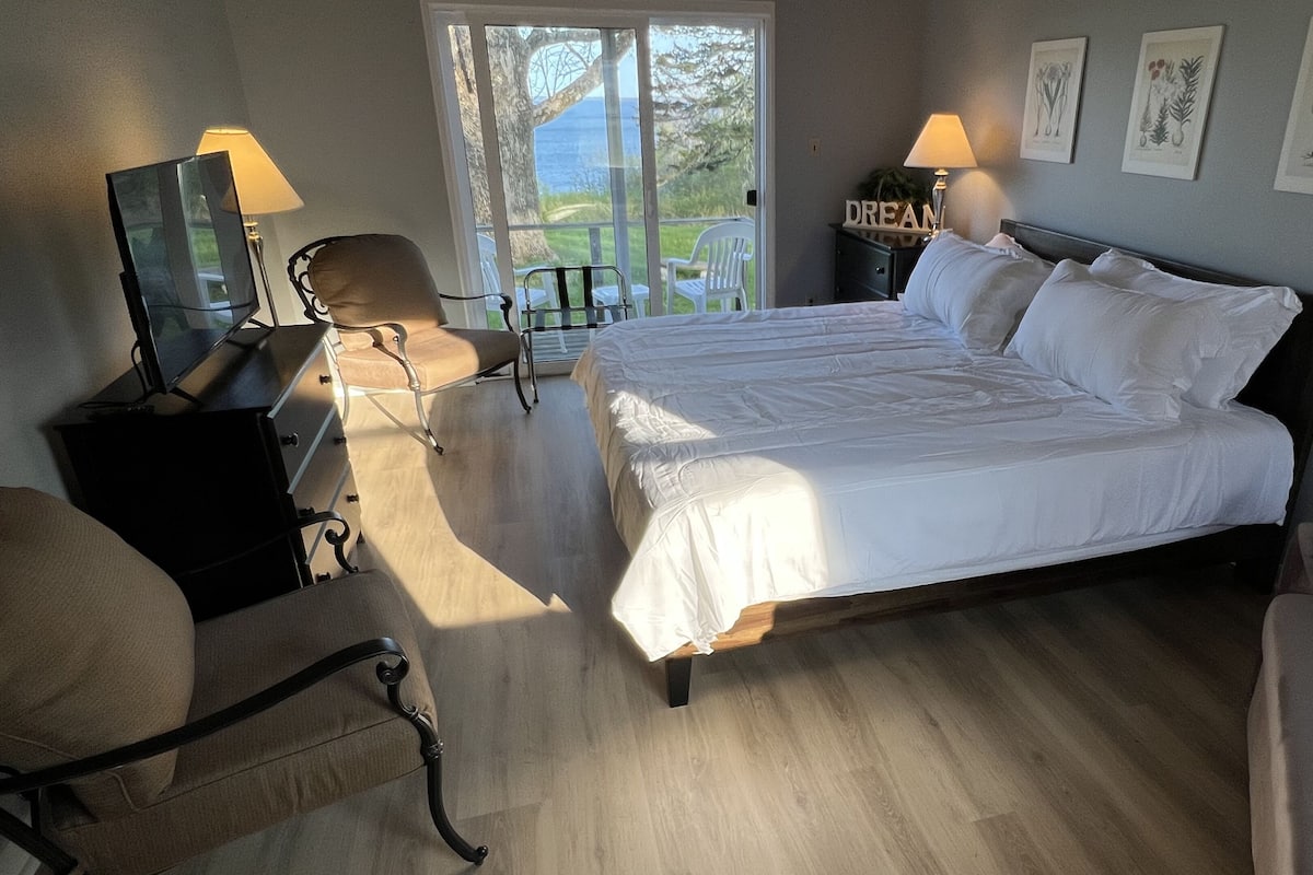 A bedroom with a large bed, two bedside tables with lamps, a chair, a desk, and a window with a view of greenery.