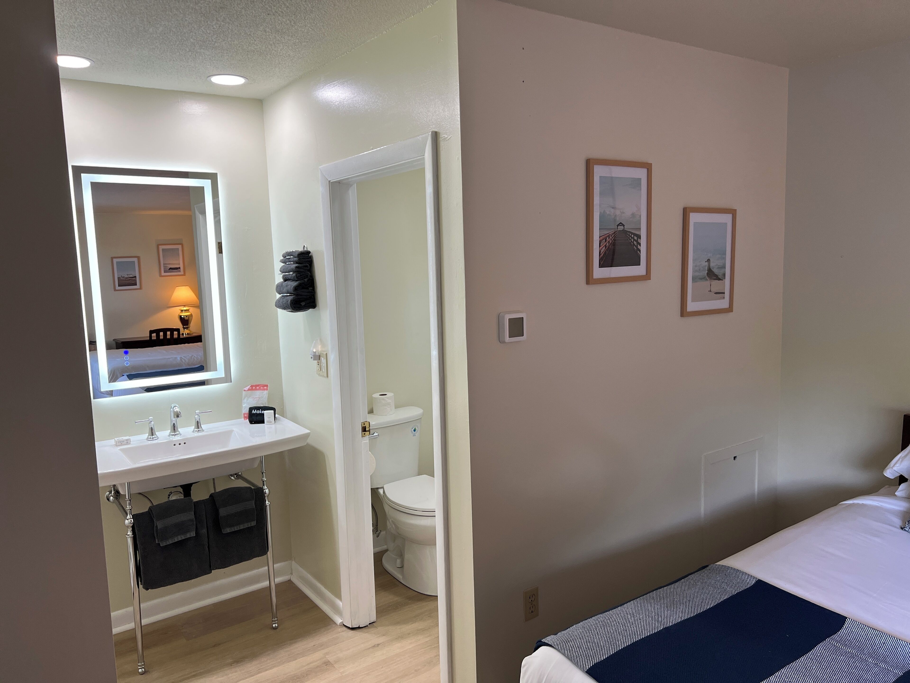 A hotel room with a bathroom featuring a sink, mirror, and toilet. There are two framed pictures on the wall.