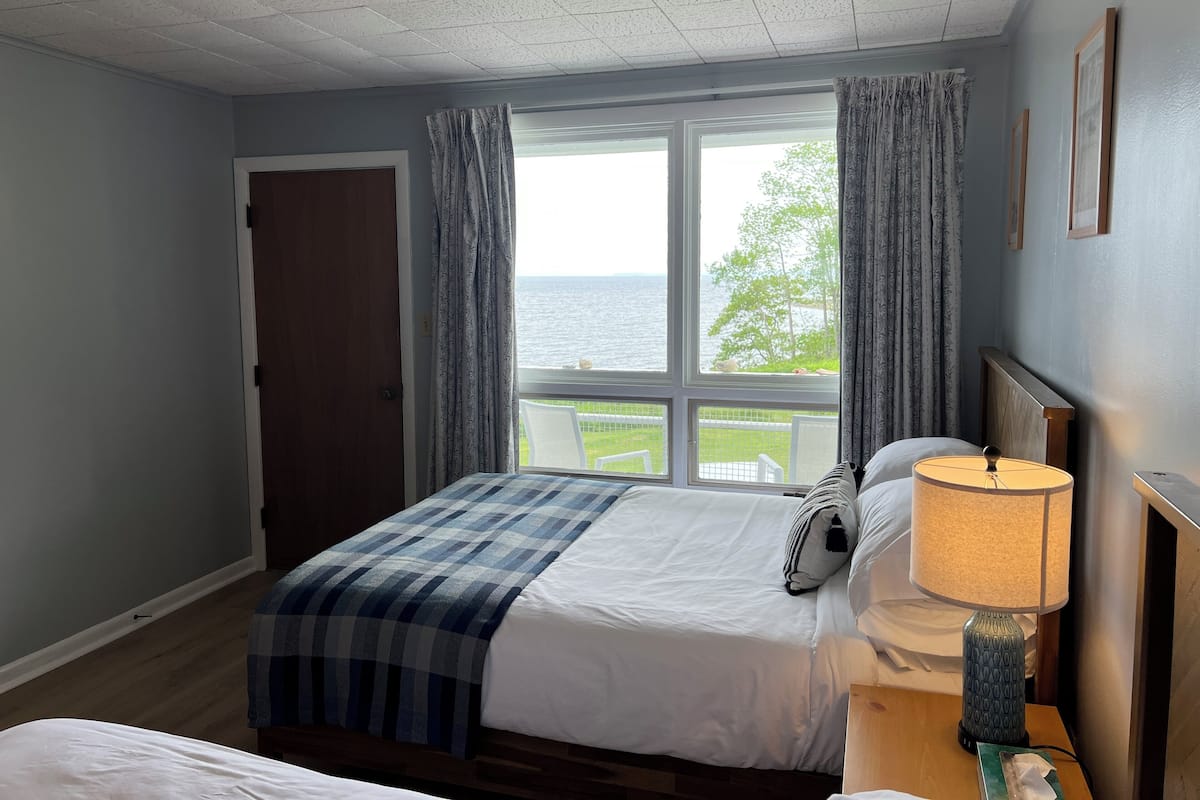 A hotel room with a large window, a bed with a blue checkered blanket, a bedside table with a lamp, and a view of the sea.