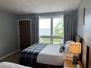 A hotel room with a large window, a bed with a blue checkered blanket, a bedside table with a lamp, and a view of the sea. - High Tide Inn (Camden)
