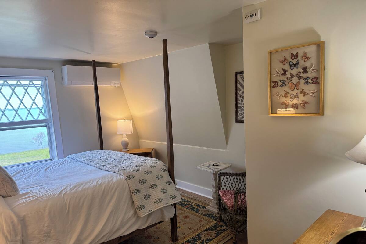 A bedroom with a bed, a nightstand, a lamp, a small table, a chair, a rug, and a framed butterfly artwork on the wall.