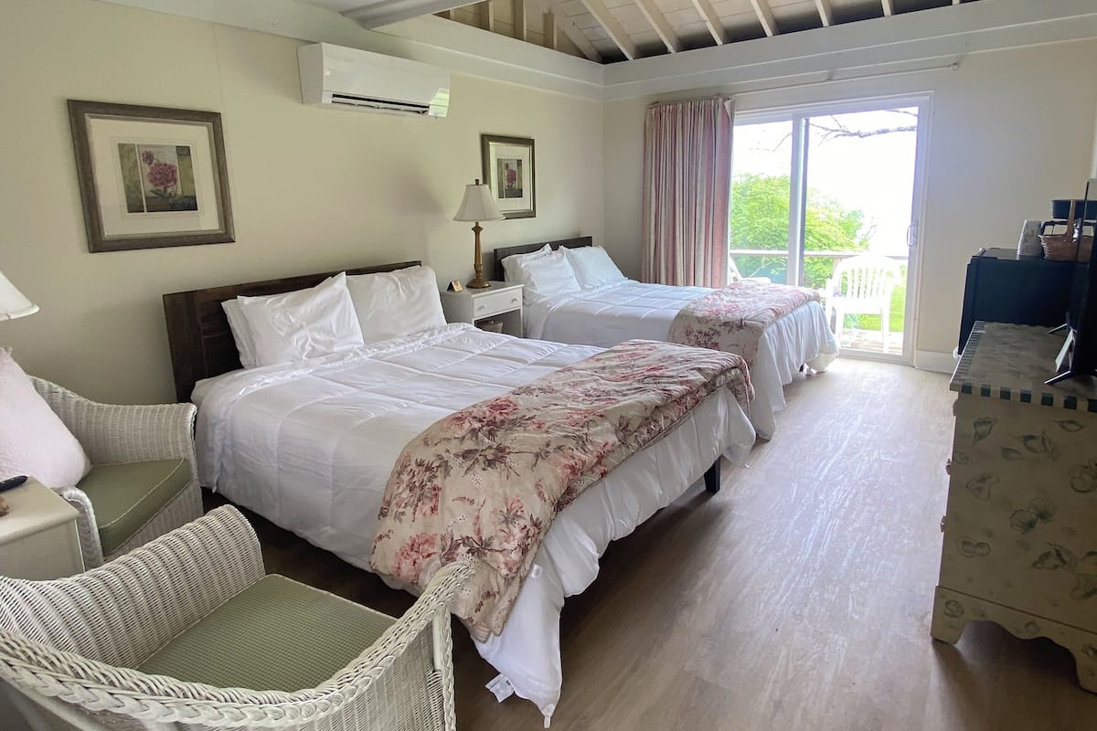 A hotel room with two beds, a wicker chair, a dresser, and a balcony with curtains.