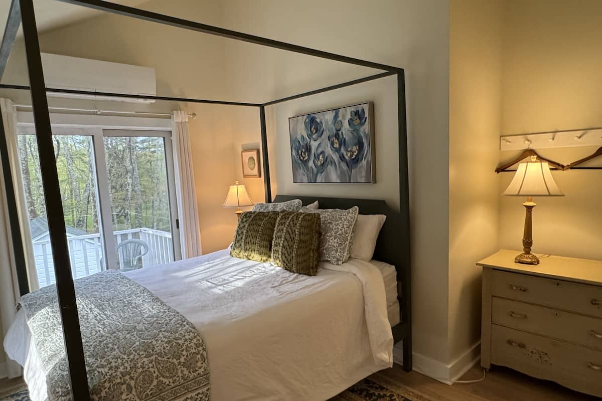 A four-poster bed with a patterned bedspread, a nightstand with a lamp, and a painting on the wall.