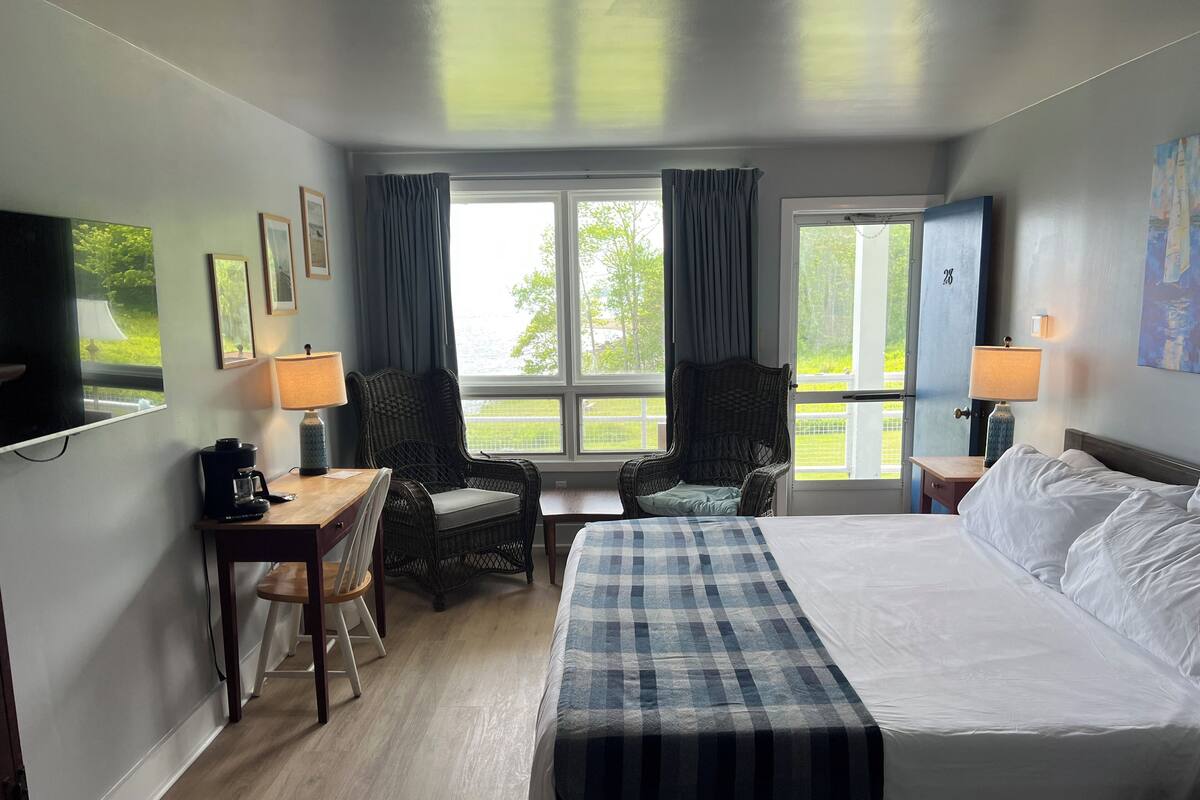 A hotel room with a large bed, a desk with a coffee maker, a wicker chair, and a view of the outdoors through the windows.