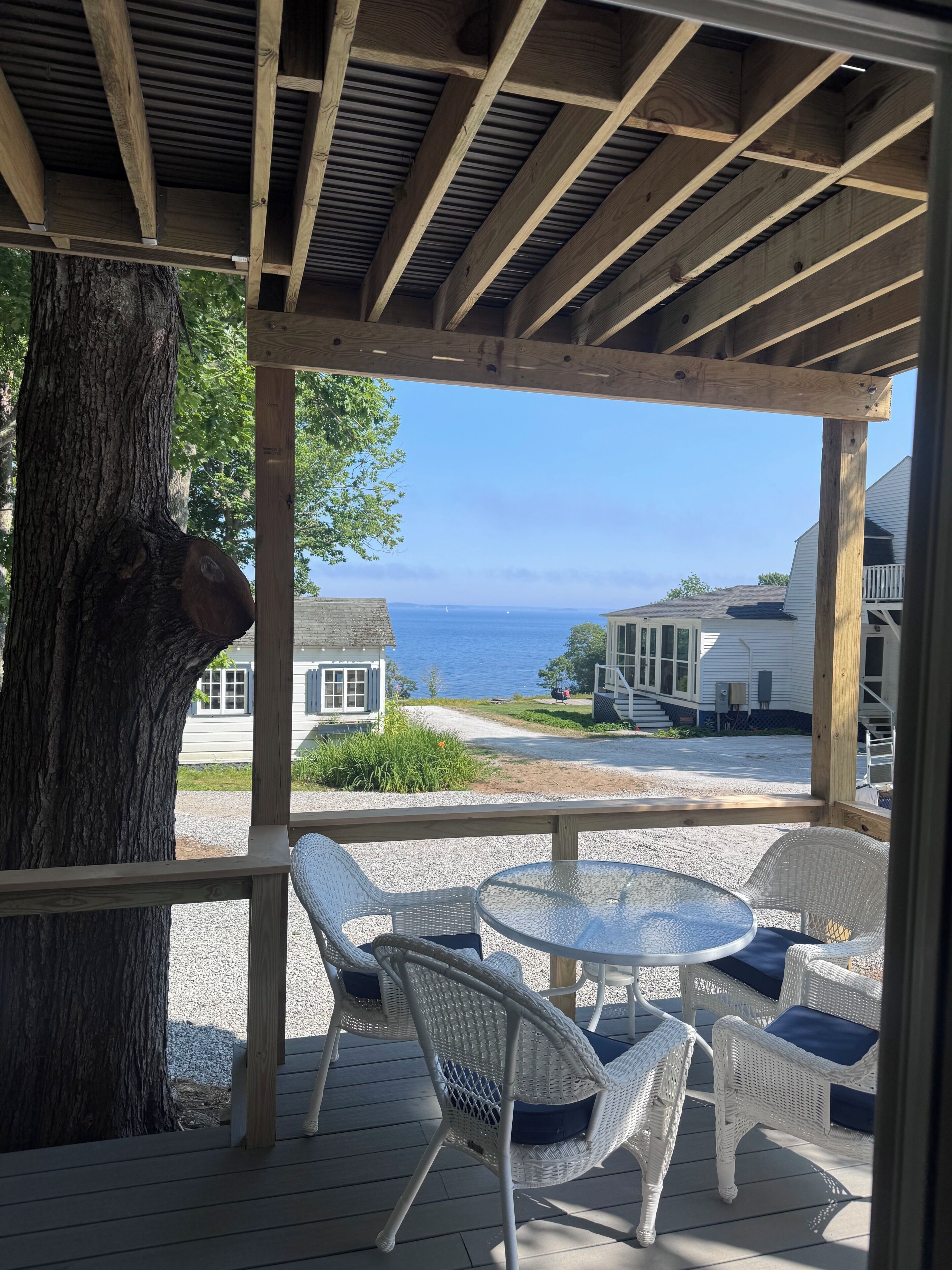 A covered outdoor seating area with wicker chairs and a table, overlooking a residential street and the sea.