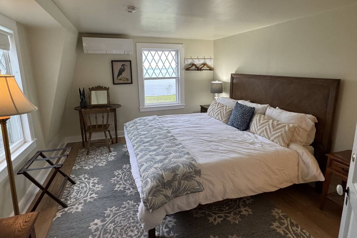 A bedroom with a large bed, a desk with a chair, a window with a view, a lamp, and a wall-mounted air conditioner.