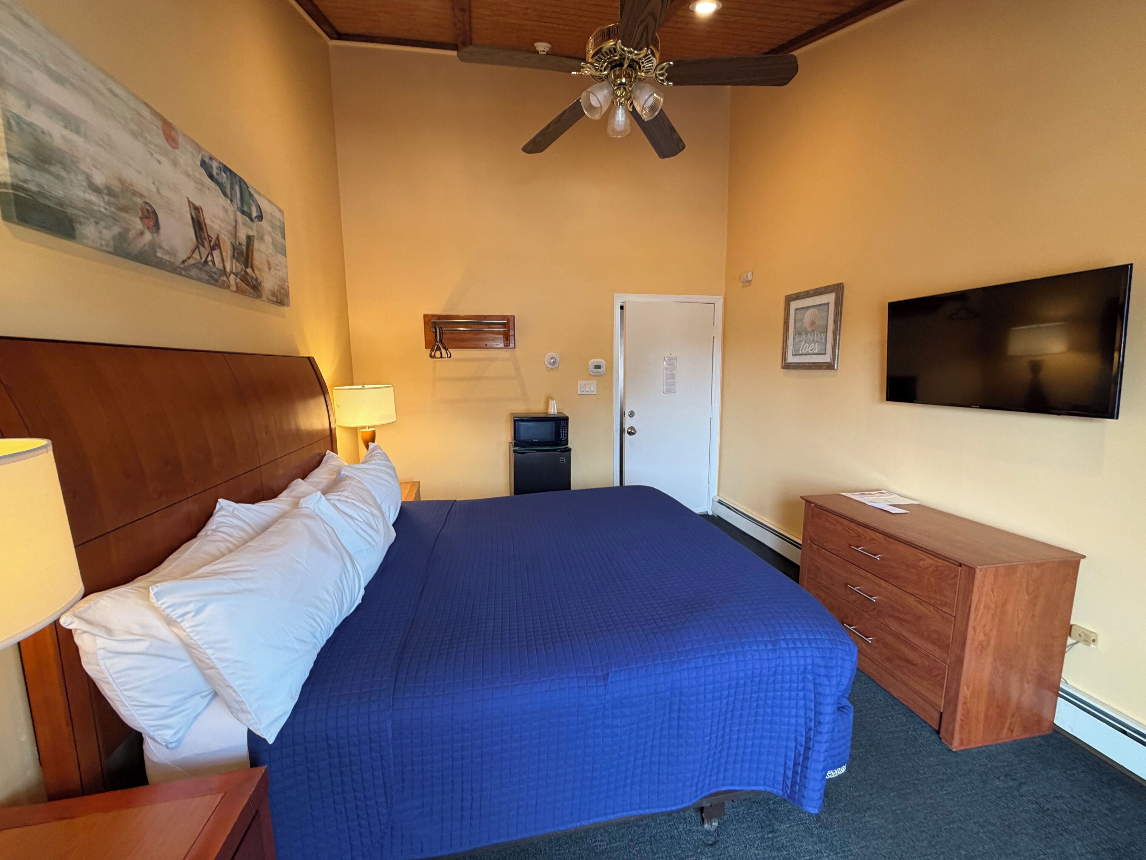A hotel room with a bed, a television, a desk, a chair, a ceiling fan, and a painting on the wall.