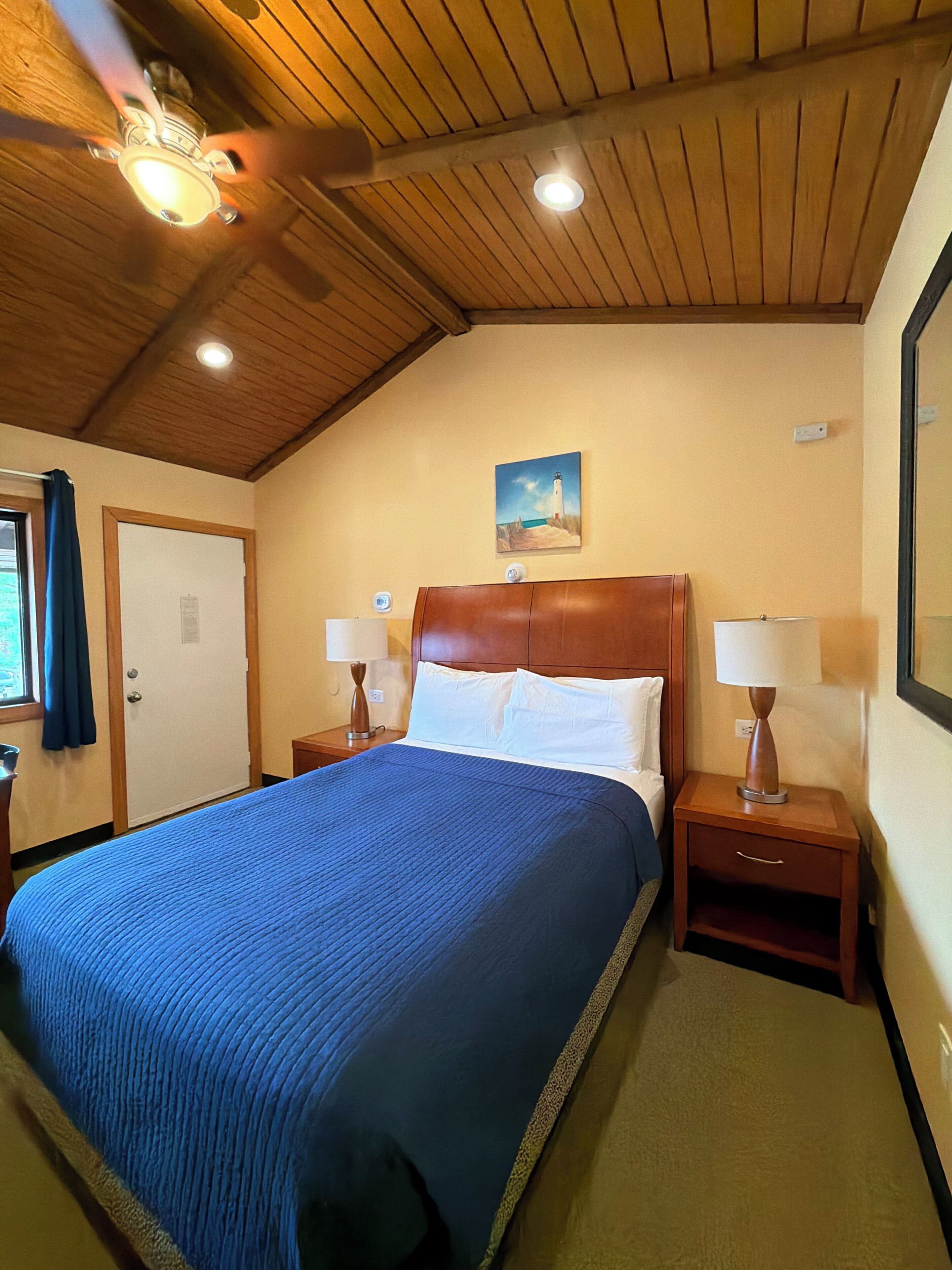 A hotel room with a large bed, wooden ceiling, two bedside tables with lamps, a painting on the wall, and a door with a curtain.