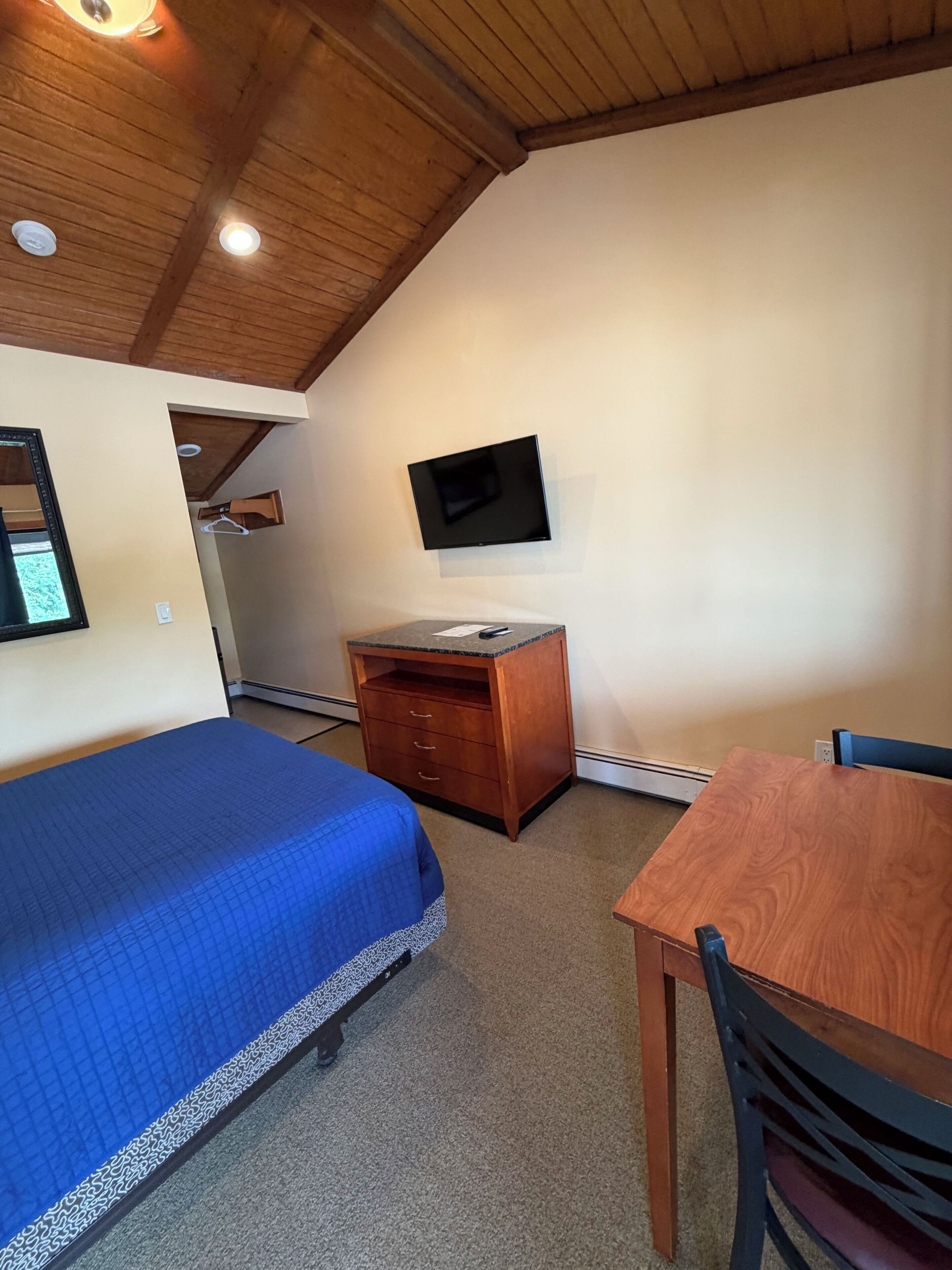 Een kamer met een bed, een bureau, een stoel, een aan de muur bevestigde televisie en een houten plafond.
