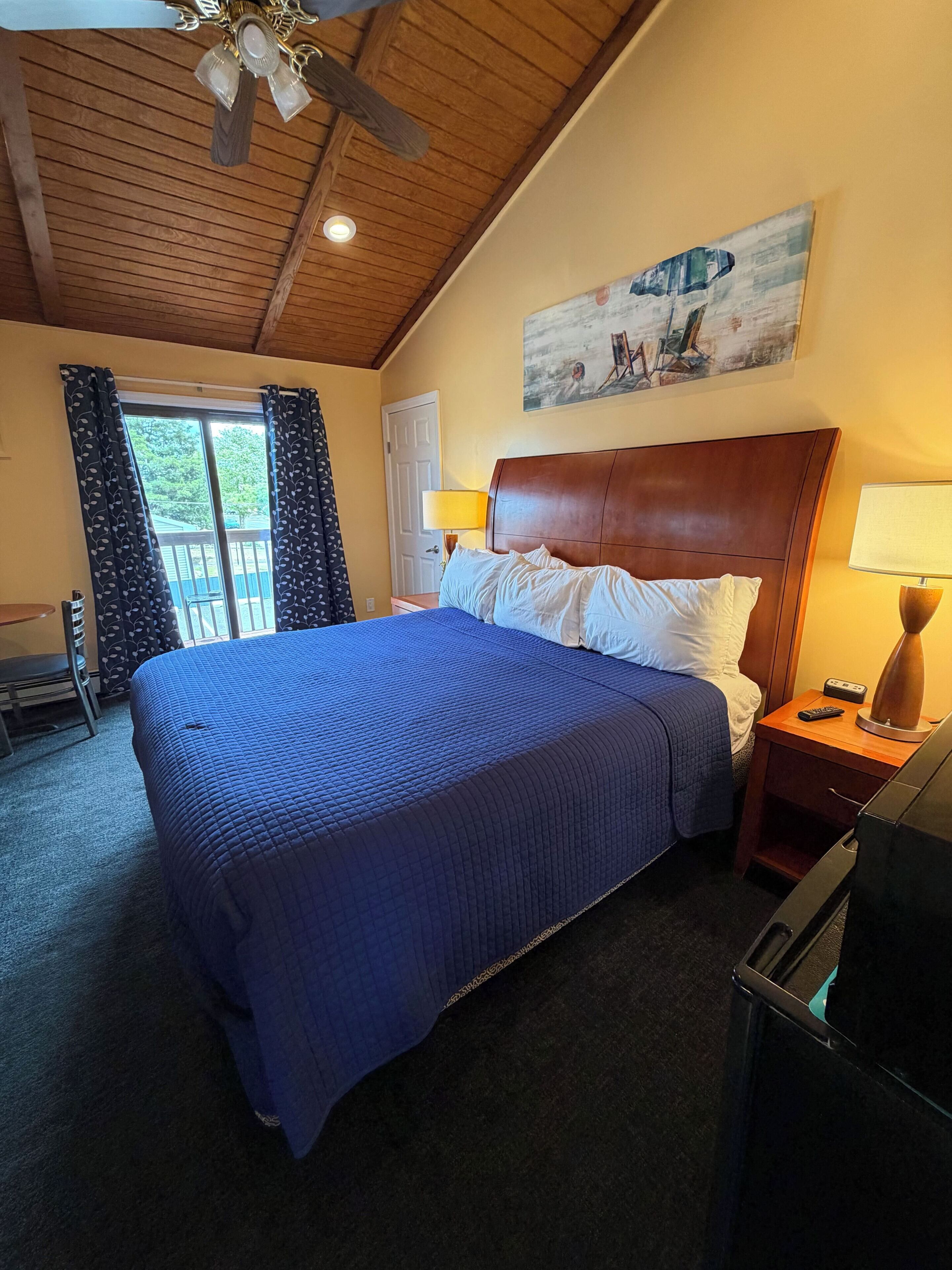 A hotel room with a bed, a wooden headboard, a painting on the wall, a window with curtains, and a ceiling fan.