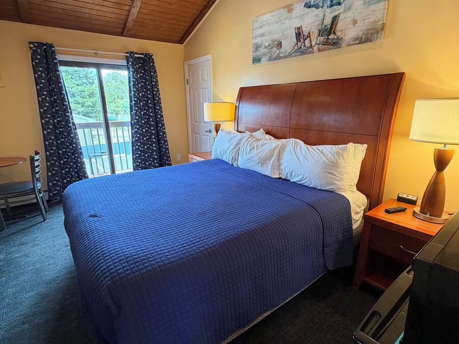 A hotel room with a bed, a wooden headboard, a painting on the wall, a window with curtains, and a ceiling fan.