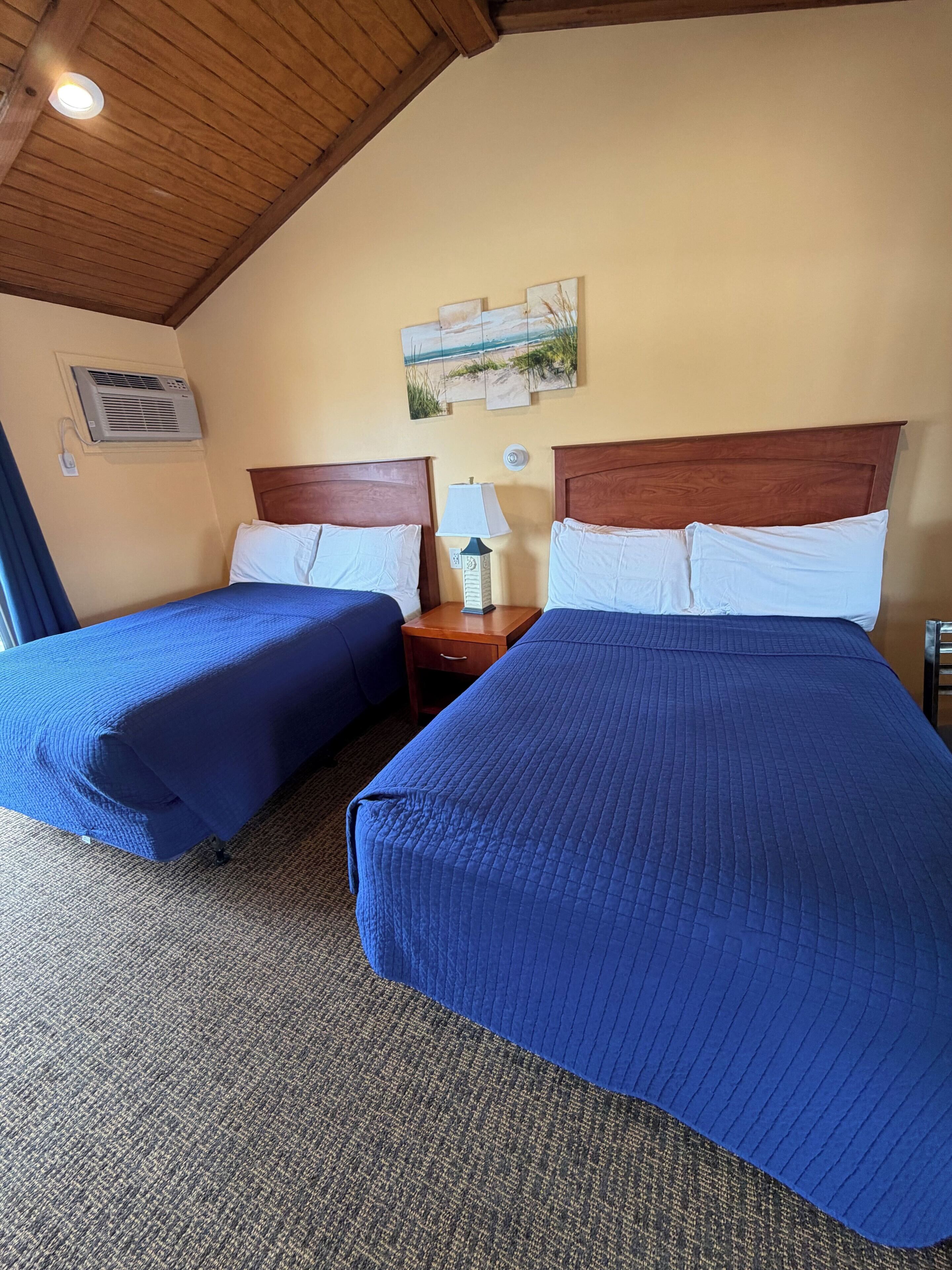 A hotel room with two beds, a wooden ceiling, a wall-mounted air conditioner, and a nightstand with a lamp.