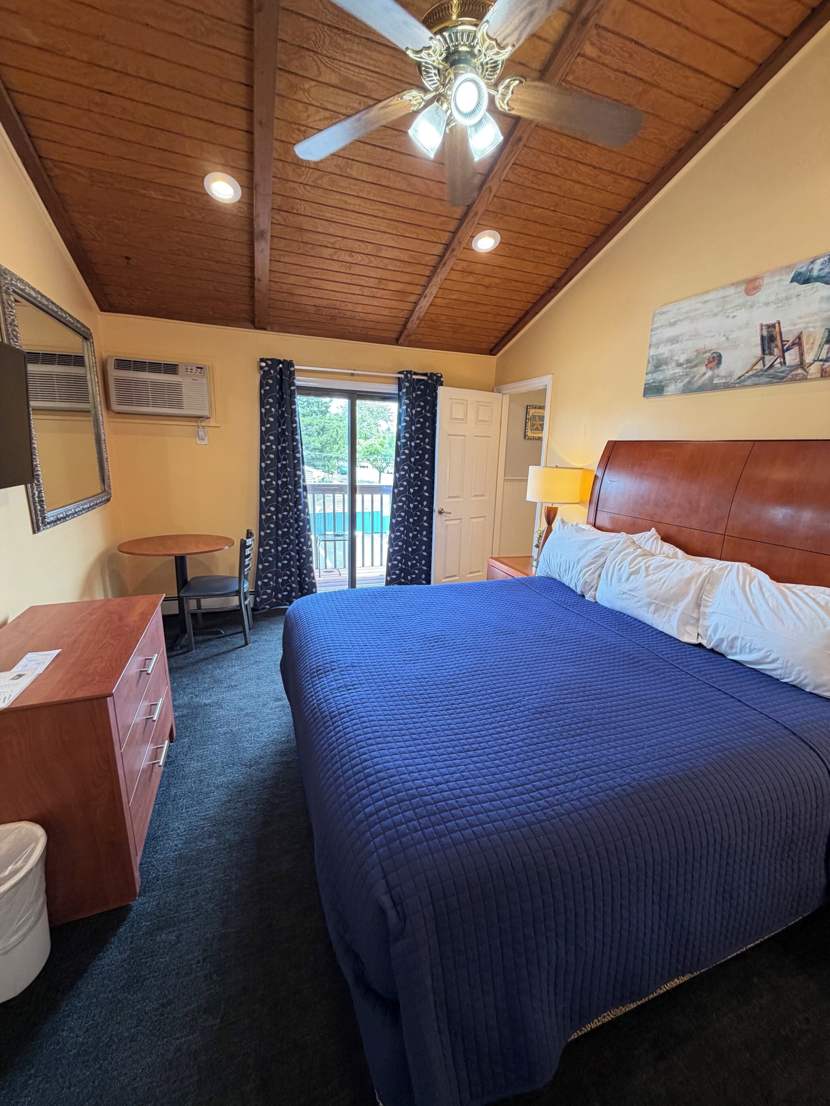 A hotel room with a bed, a desk, a chair, a ceiling fan, and a painting on the wall.