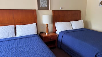 A hotel room with two beds, each with a blue quilted bedspread, a wooden headboard, and white pillows. There is a nightstand with a lamp between the beds.