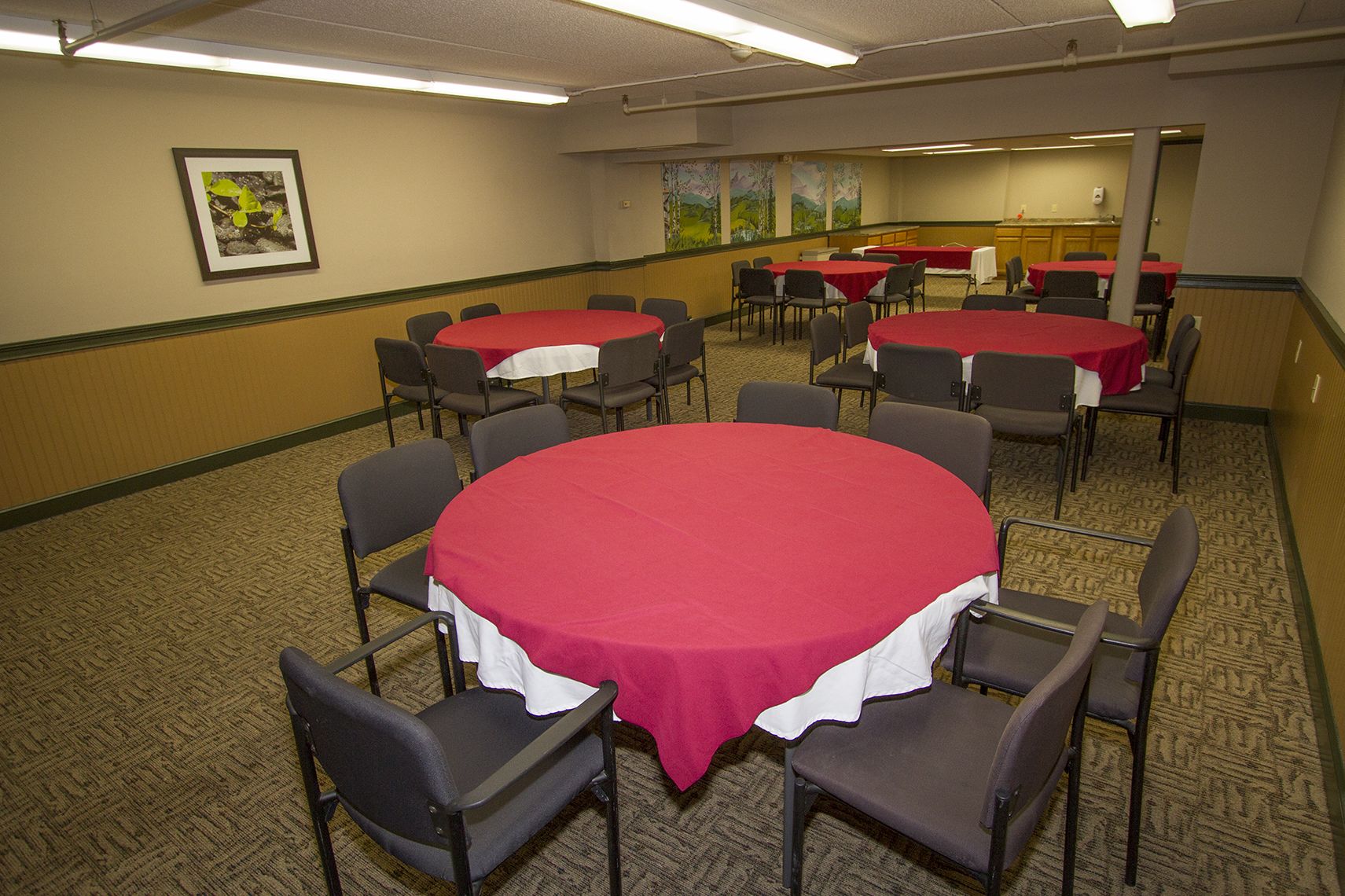 A conference room with round tables covered in red tablecloths, surrounded by chairs, and a kitchen area in the background.