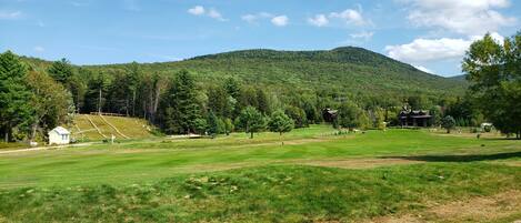 A golf course with a clubhouse, a small white building, and a hill in the background.