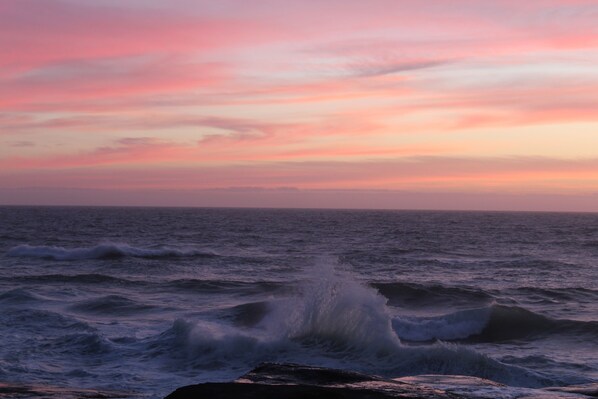 A coastal scene with waves crashing onto rocks under a pink and purple sky. - Overleaf Lodge (Yachats)