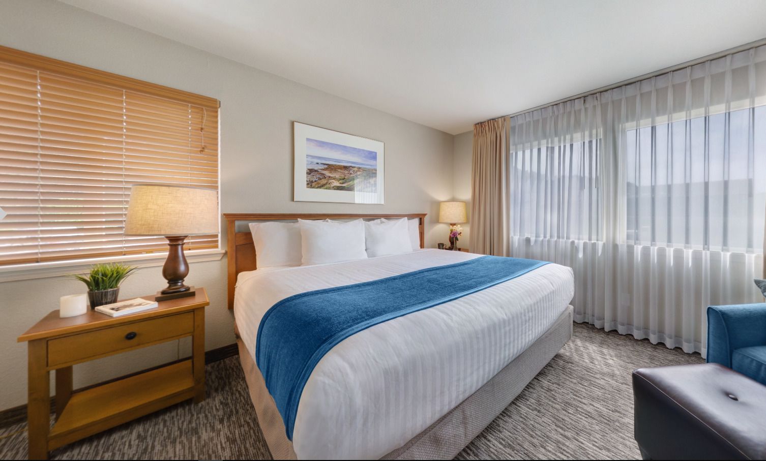 A hotel room with a large bed, a wooden nightstand with a lamp, a small table with a plant, a blue throw blanket, and a view of the outdoors through sheer curtains.