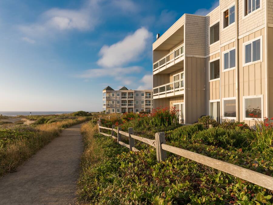 A coastal building with multiple balconies, a paved pathway, and surrounding vegetation.