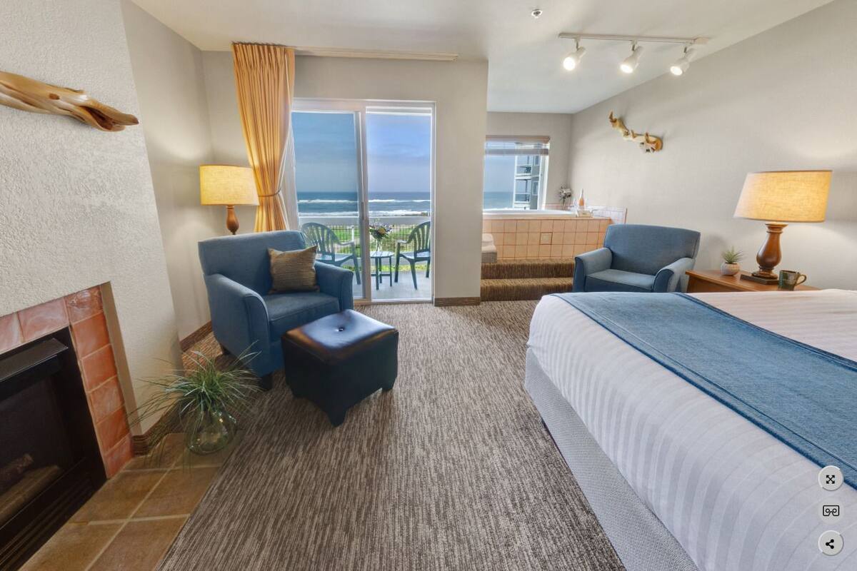 A hotel room with a fireplace, a bed, two armchairs, a small table, and a view of the ocean through a sliding door.