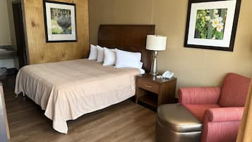 A hotel room with a large bed, a wooden headboard, a nightstand with a lamp and phone, a red chair, and two framed pictures on the walls.