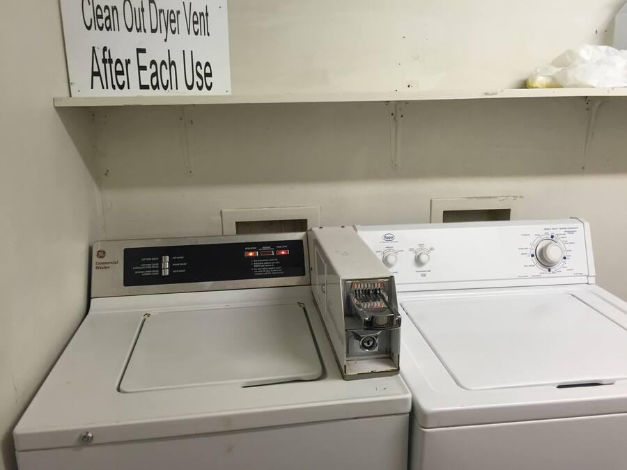 Two front-loading washers with control panels, a sign instructing to clean the dryer vent after each use, and a shelf above the dryers.