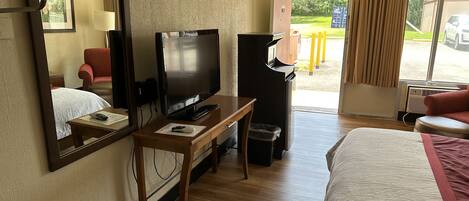 Ett hotellrum med en säng, en tv, ett skrivbord, en spegel och ett fönster med gardiner.