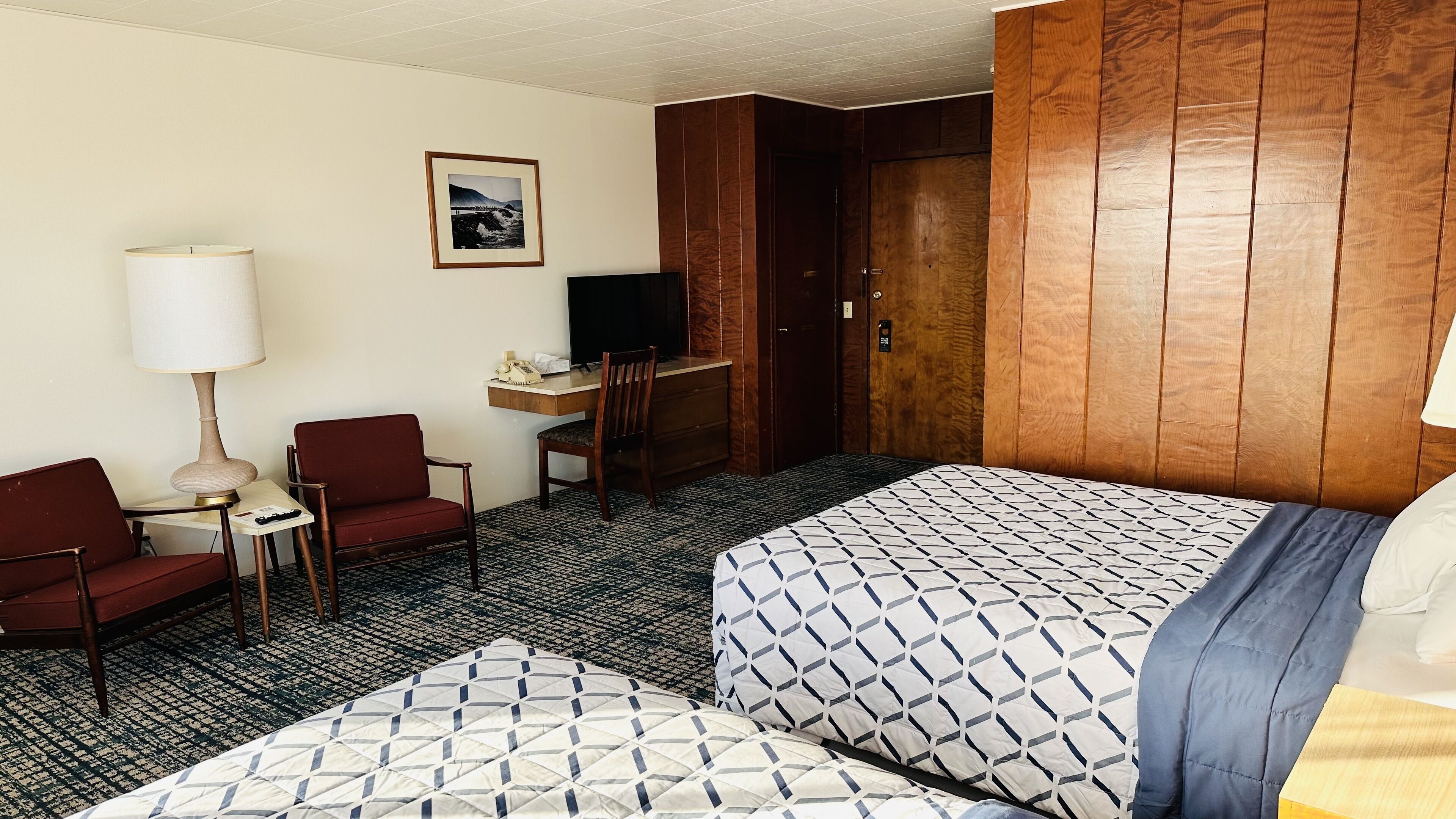 A hotel room with two beds, a desk, a chair, a TV, and a lamp.