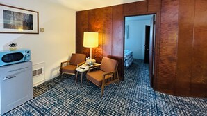 A hotel room with a wooden panel wall, a small desk with a lamp, two chairs, a microwave, and a bed with a checkered blanket. - Curly Redwood Lodge (Crescent City)