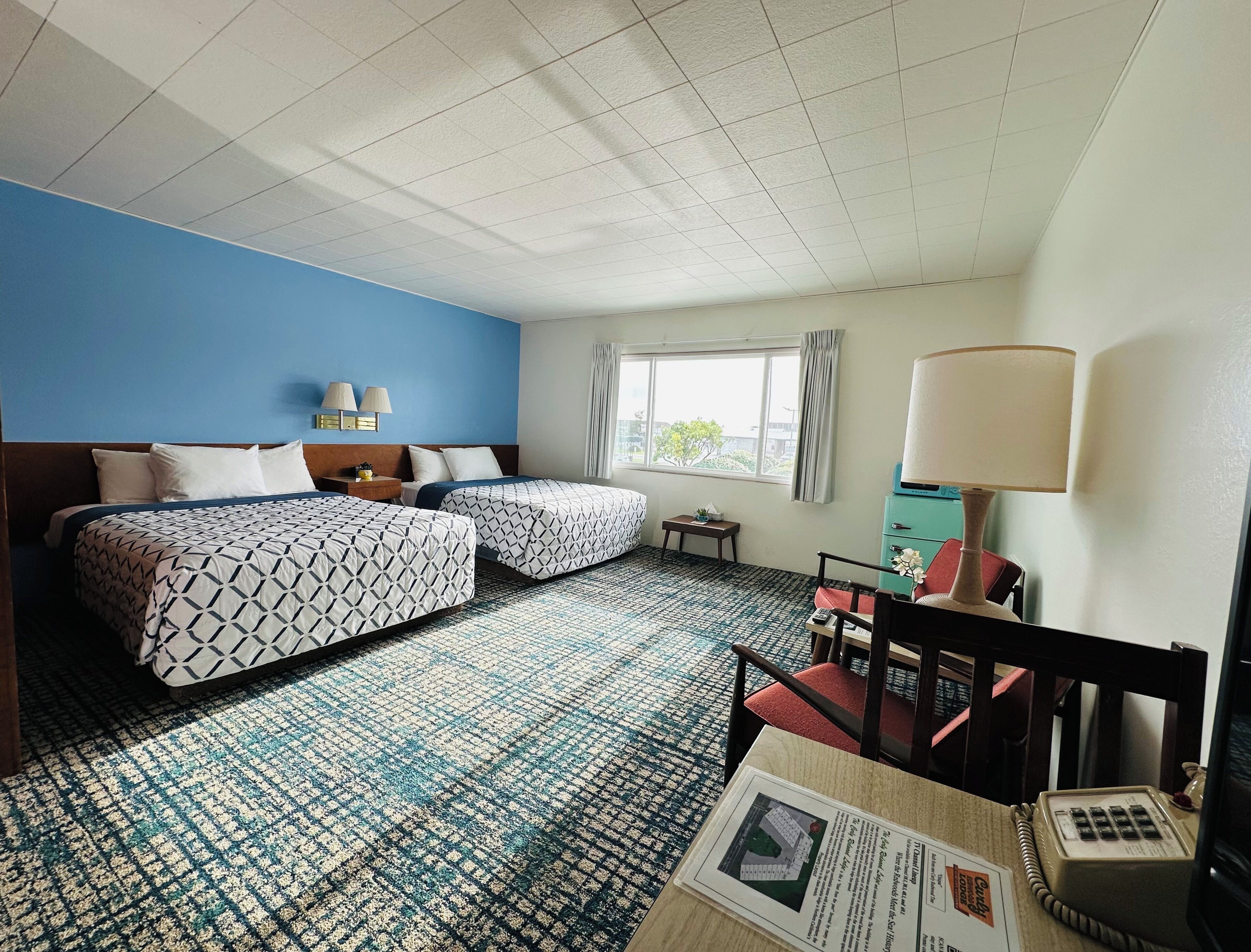 Quarto de hotel com duas camas, uma mesinha, uma cadeira, um abajur e uma janela com cortinas.