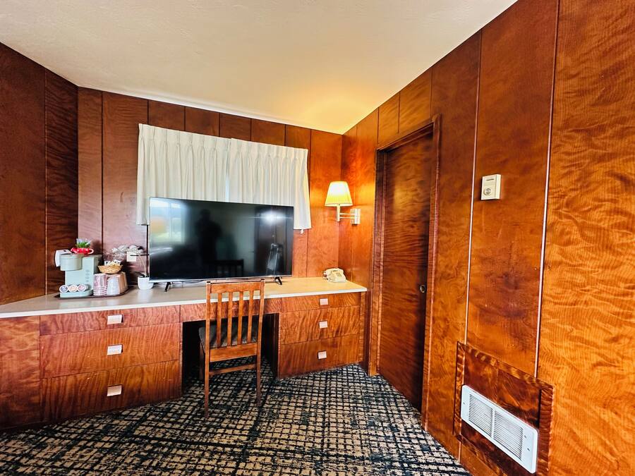 A hotel room with a wooden paneled wall, a large flat-screen TV, a desk with a chair, a lamp, and a telephone.