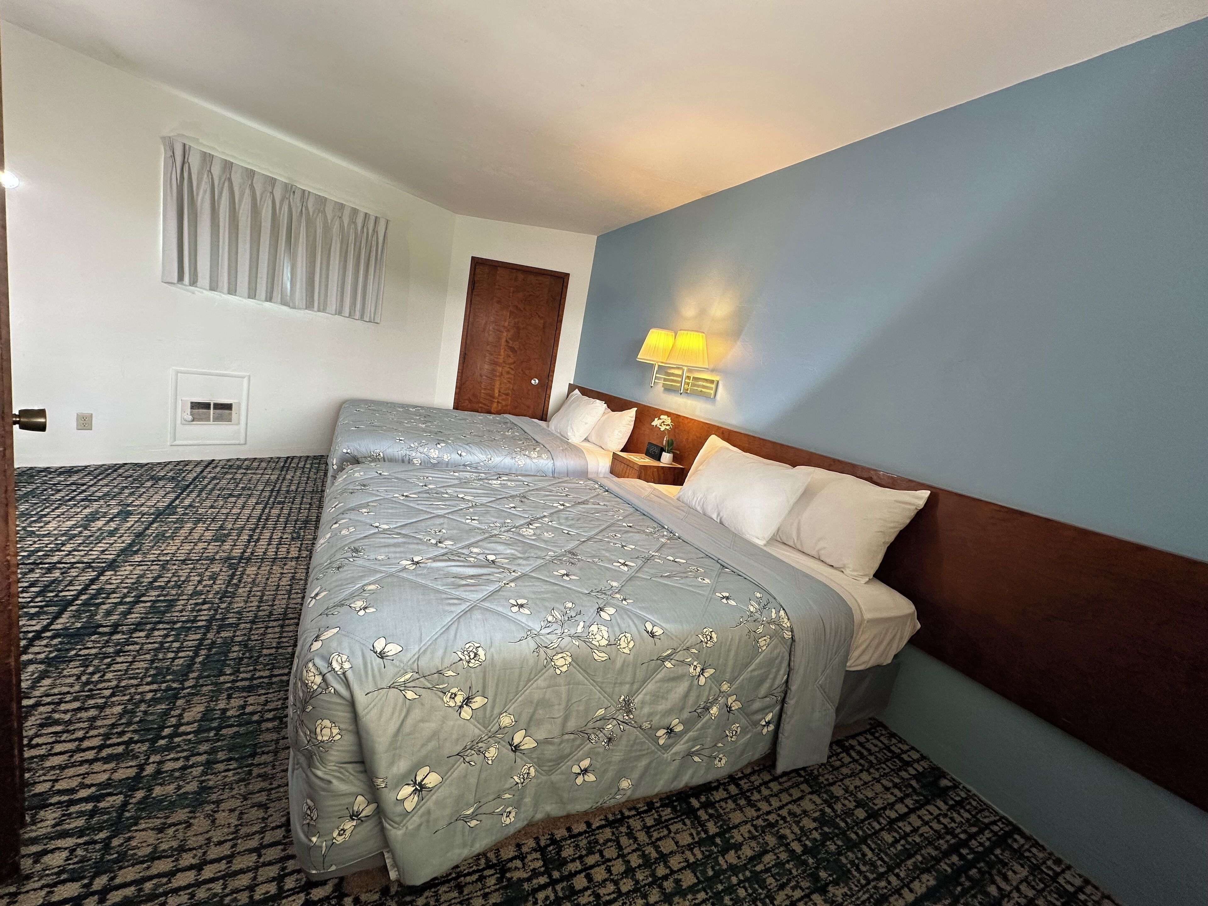 A hotel room with a bed, a nightstand, a lamp, a window with curtains, and a wall-mounted air vent.