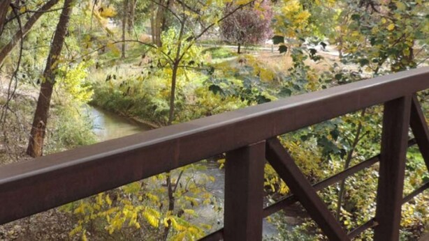 Un puente de madera con barandilla de metal que da vista a un paisaje natural con árboles y un pequeño arroyo.