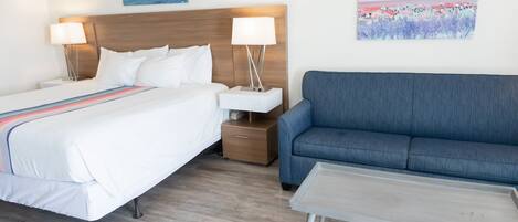 Hotellihuone, jossa on sänky, sohva, yöpöytä, lamppu ja kaksi taulua seinällä.