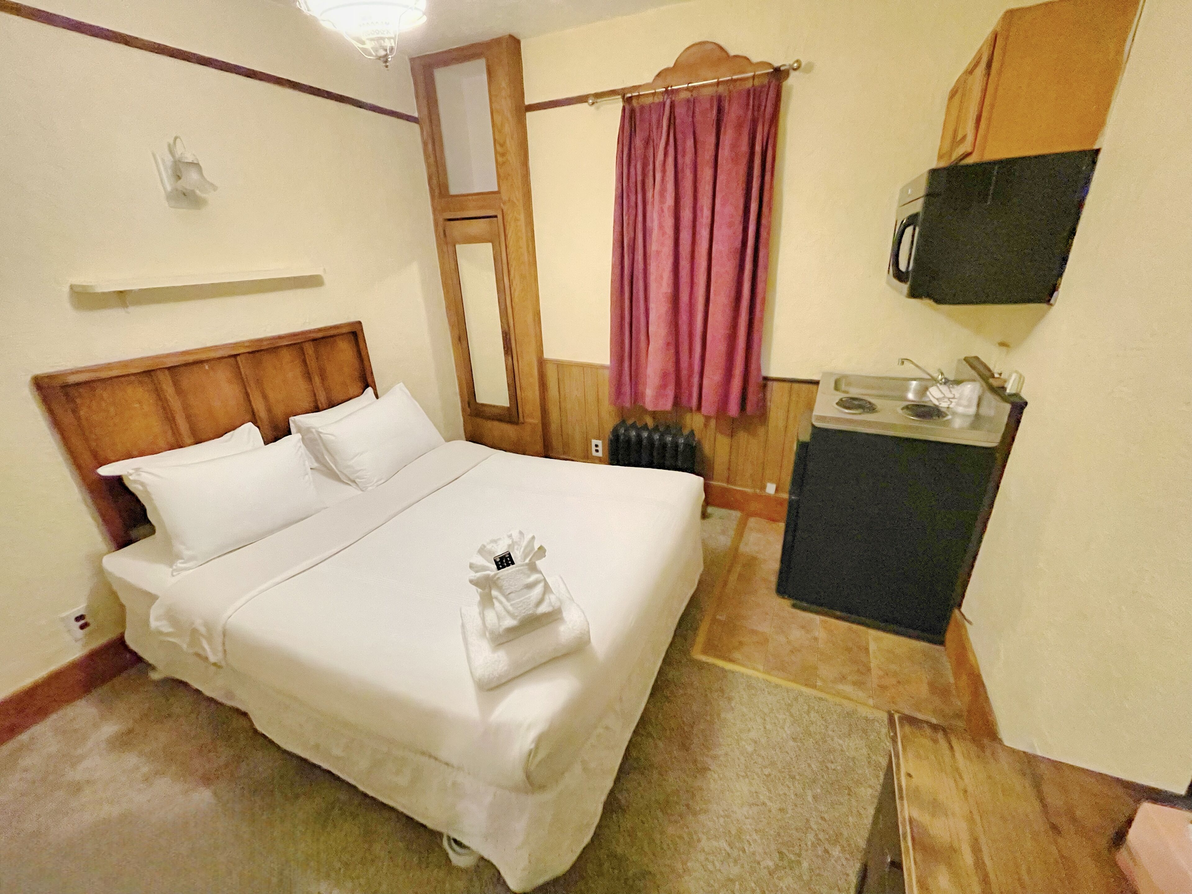 Habitación de hotel con cama, kitchenette pequeña, espejo, ventana con cortina y televisor montado en la pared.