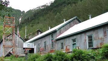 An old, weathered building with a metal staircase and a signboard, surrounded by dense greenery and mountains.