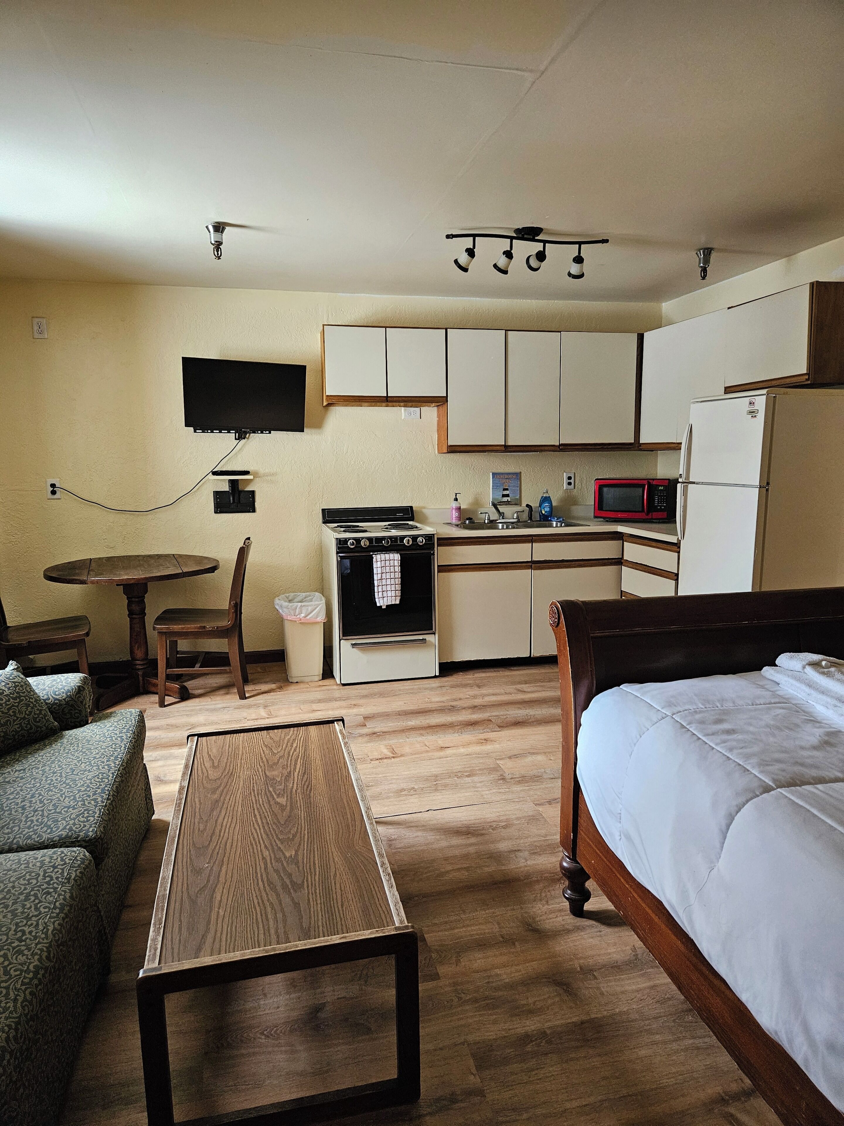 A compact room with a bed, a wooden table, a kitchenette with a stove, microwave, and refrigerator, a TV mounted on the wall, and a small round table with chairs.