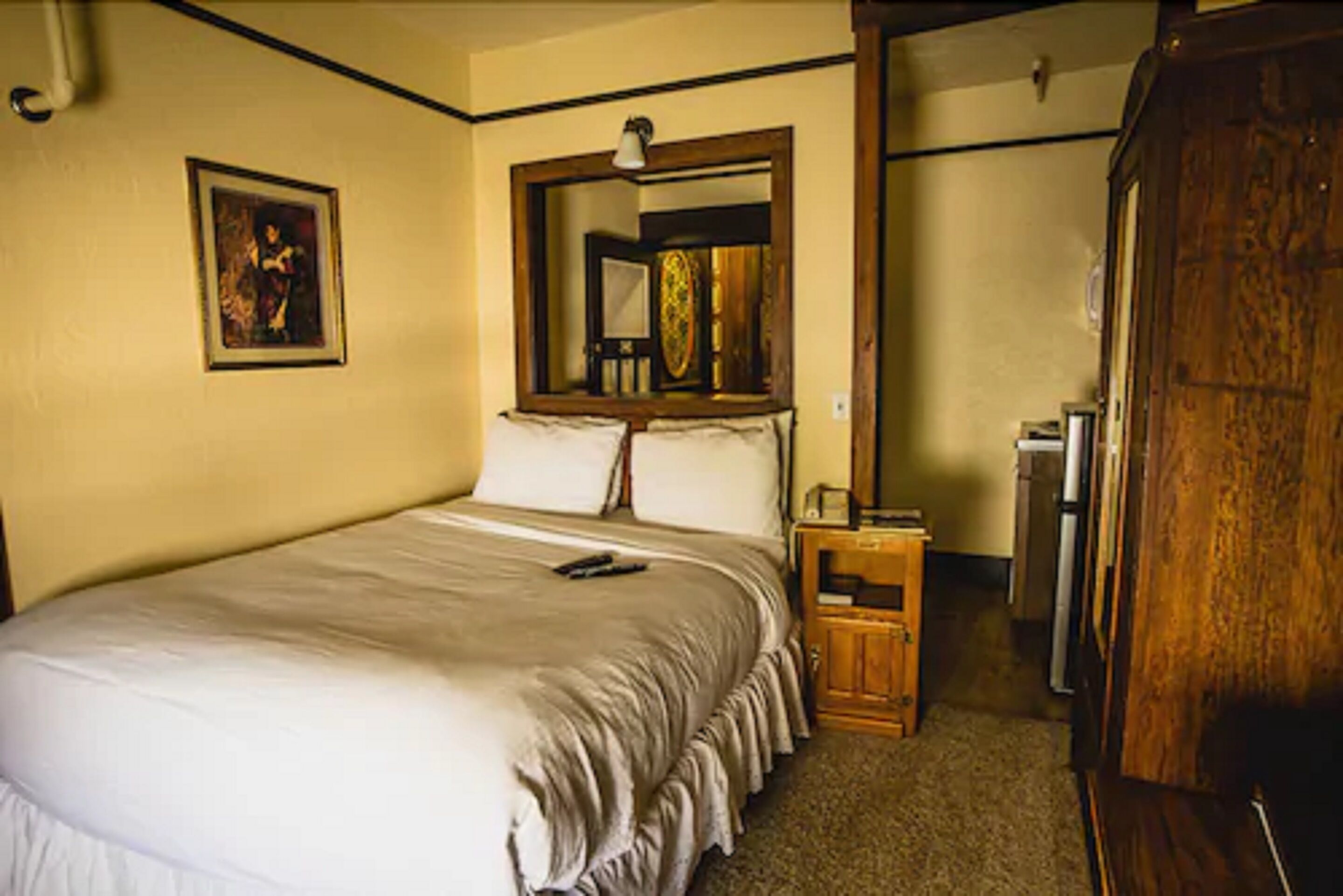 Habitación de hotel con una cama grande, un armario de madera, una mesita de noche pequeña, un espejo y un cuadro en la pared.