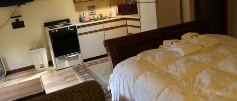 A hotel room with a bed, a wooden headboard, a kitchenette with a stove, microwave, and refrigerator, a small rug, and a television mounted on the wall.
