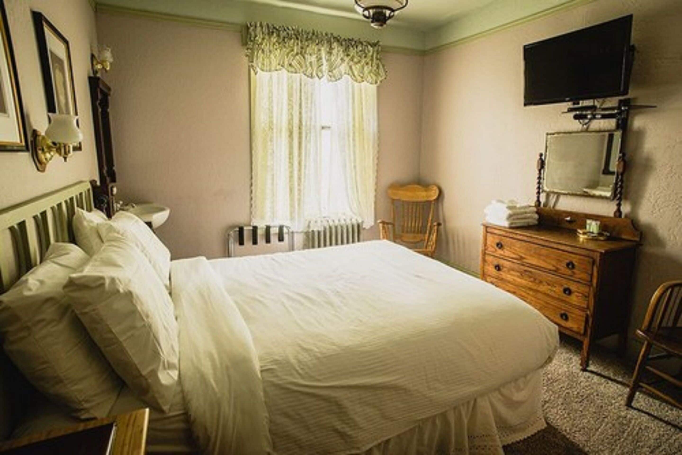 A bedroom with a large bed, a dresser, a chair, a radiator, a television, and a window with curtains.