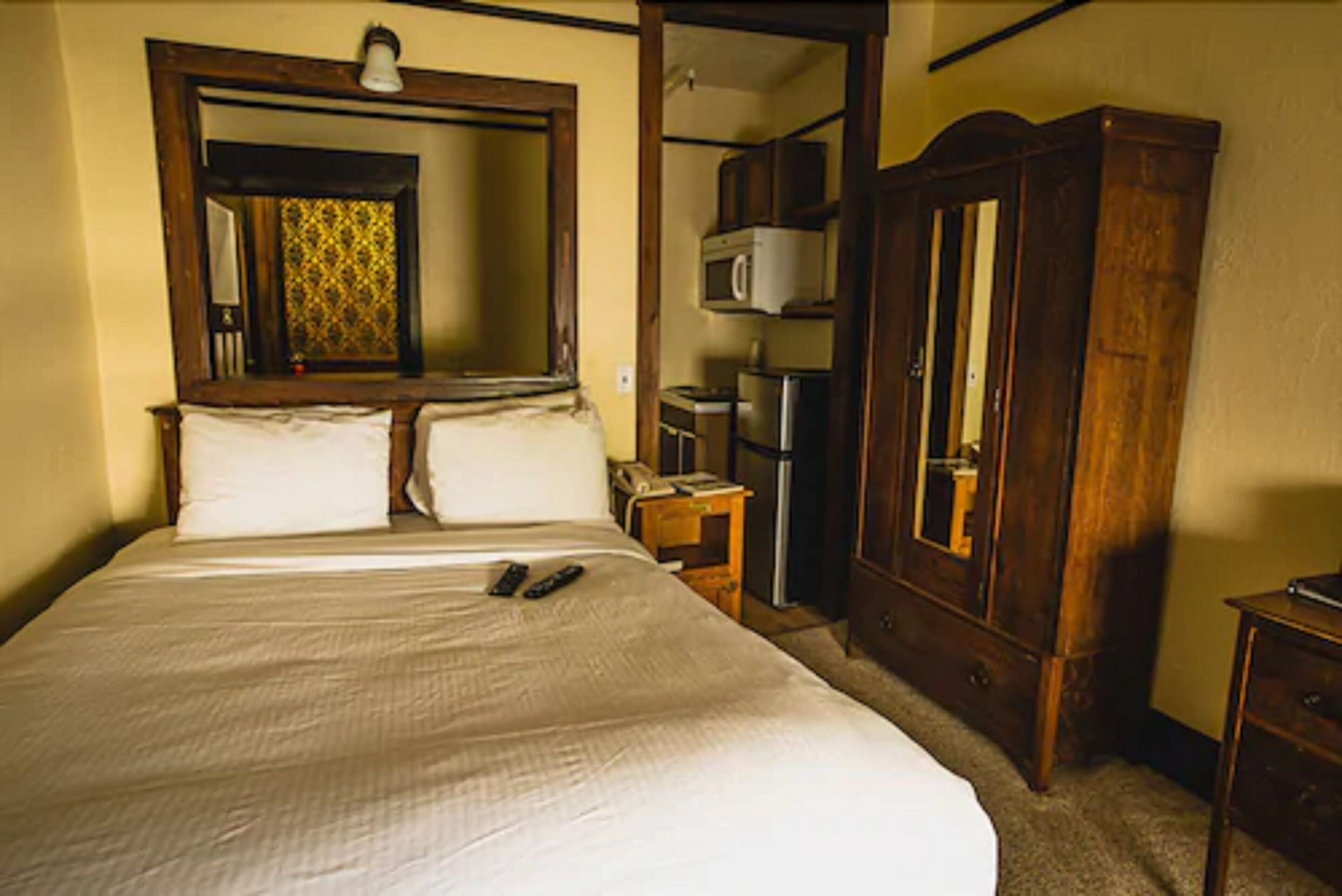 Quarto com uma cama grande, um guarda-roupa de madeira, uma pequena escrivaninha com uma lâmpada e um espelho sobre a cama.